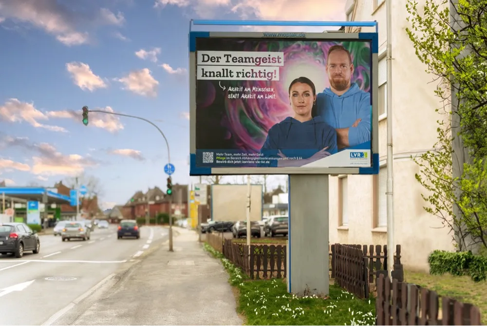 Foto eines Großflächenplakates als Out-Of-Home Maßnahme, auf dem ein Kampagnenmotiv mit einer Nahaufnahme einer selbstbewusst lächelnden Pflegerin und Pflegers vor einem abstrakten Hintergrund mit Lavalampen-Effekten. Text auf dem Bild: 'Der Teamgeist knallt richtig! Arbeit am Menschen, statt Arbeit am Limit. LVR-Klinik Viersen' zu sehen ist.
