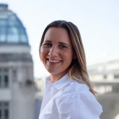 Porträt einer lächelnden Frau im weißen Hemd vor urbanem Hintergrund mit Stadtgebäuden.