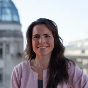 Annette Raschke mit rosa Blazer lächelt herzlich, im Hintergrund eine Dachterrasse mit urbaner Skyline.